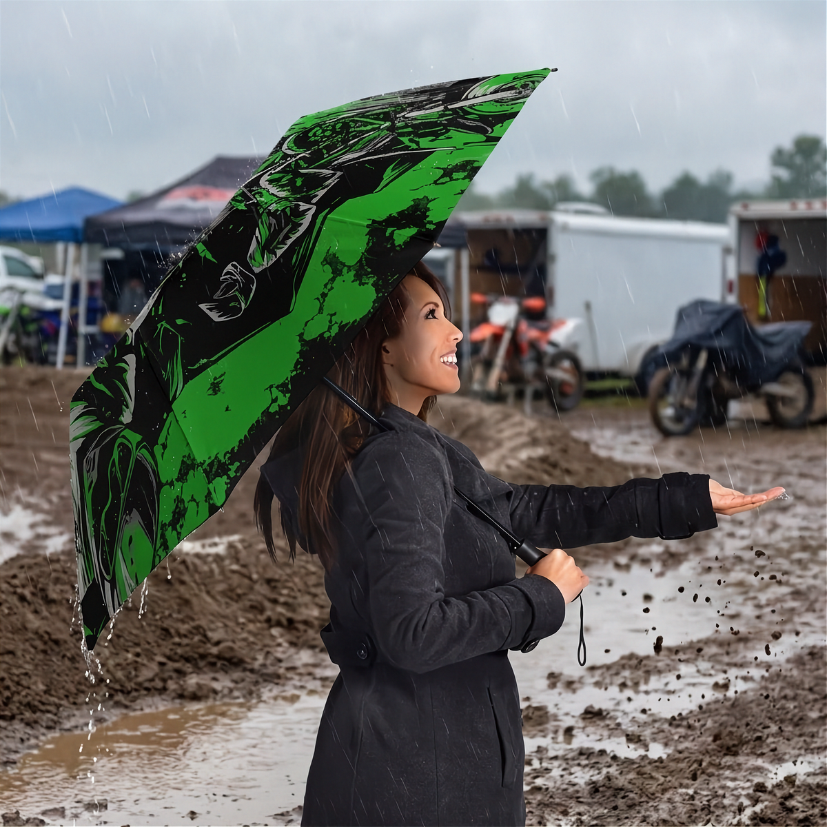 A woman in a black raincoat stands in the rain holding a large green and black printed umbrella, with muddy ground and trailers in the background.