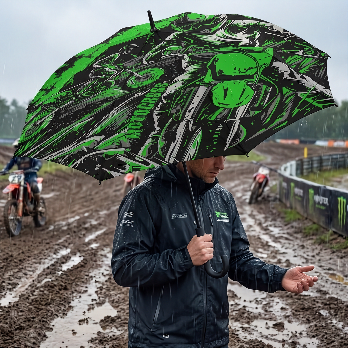 A person in a black rain jacket stands in the rain on a muddy motocross track holding a large black umbrella with a bright green and white graphic print of a motorcycle.