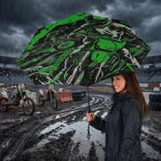 A woman in a black raincoat holds a large open umbrella with a green and black motorcycle graphic print, standing in the rain at a motocross track with dirt bikes and muddy tracks in the background.