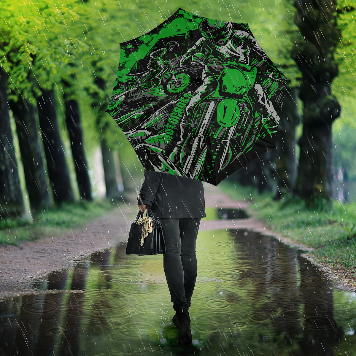 A person standing in a rain puddle on a tree-lined path, holding an open umbrella with a bold green, black, and white graphic print featuring a motorcycle and rider.