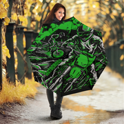 A woman standing on a wet path in an autumn park holds up a large open umbrella. The umbrella features a bold graphic print of a motorcyclist in black, white, and bright green.