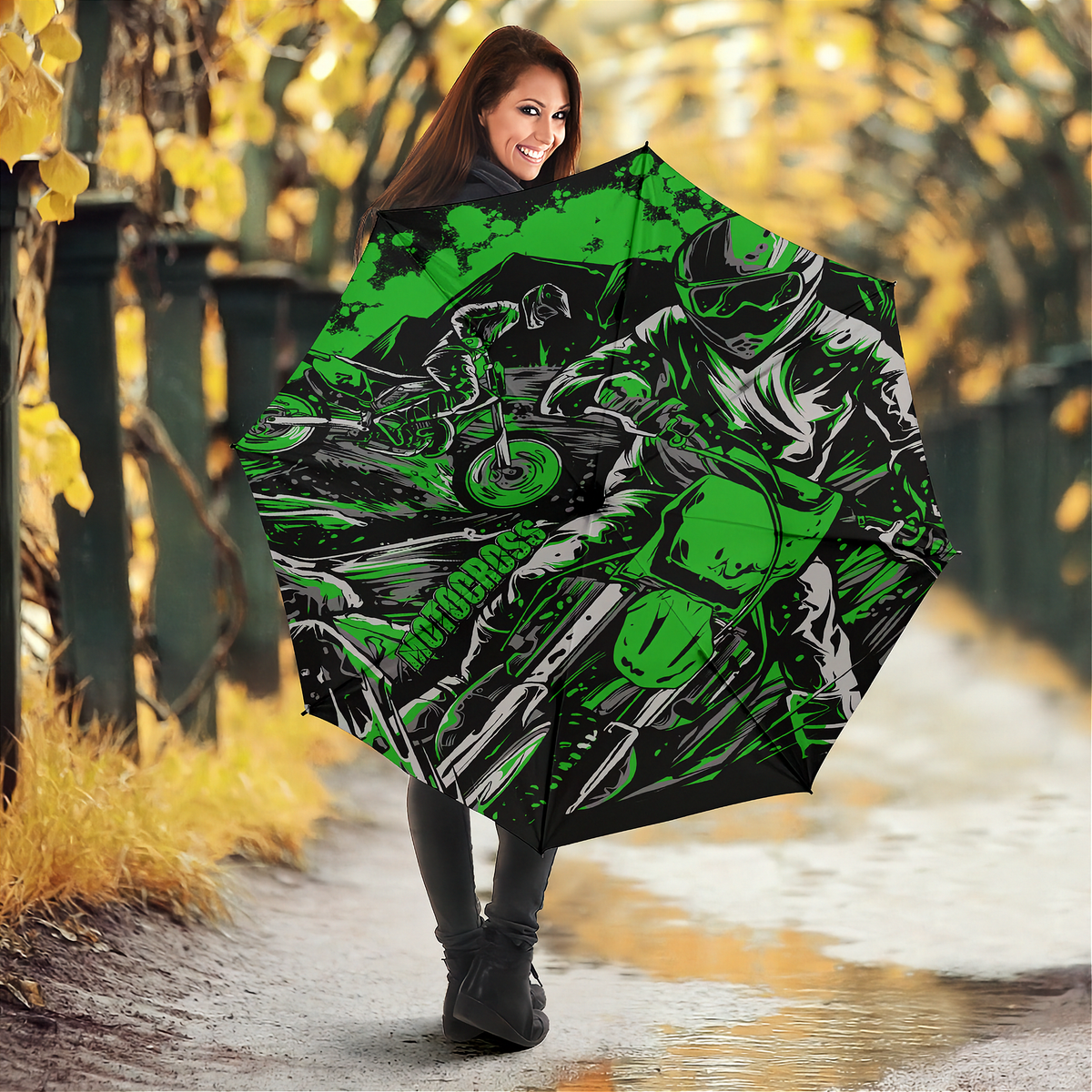 A woman standing on a wet path in an autumn park holds up a large open umbrella. The umbrella features a bold graphic print of a motorcyclist in black, white, and bright green.