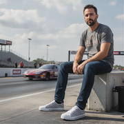 Man wearing checkered shoes sitting on a concrete block at a race track with a car in the background