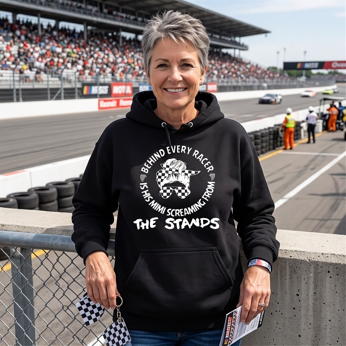A smiling woman stands at a racetrack wearing a black hoodie with a white graphic that reads 'BEHIND EVERY RACER is his MIMI SCREAMING FROM THE STANDS' with a checkered flag illustration. The background shows a race track, grandstands, and race cars.