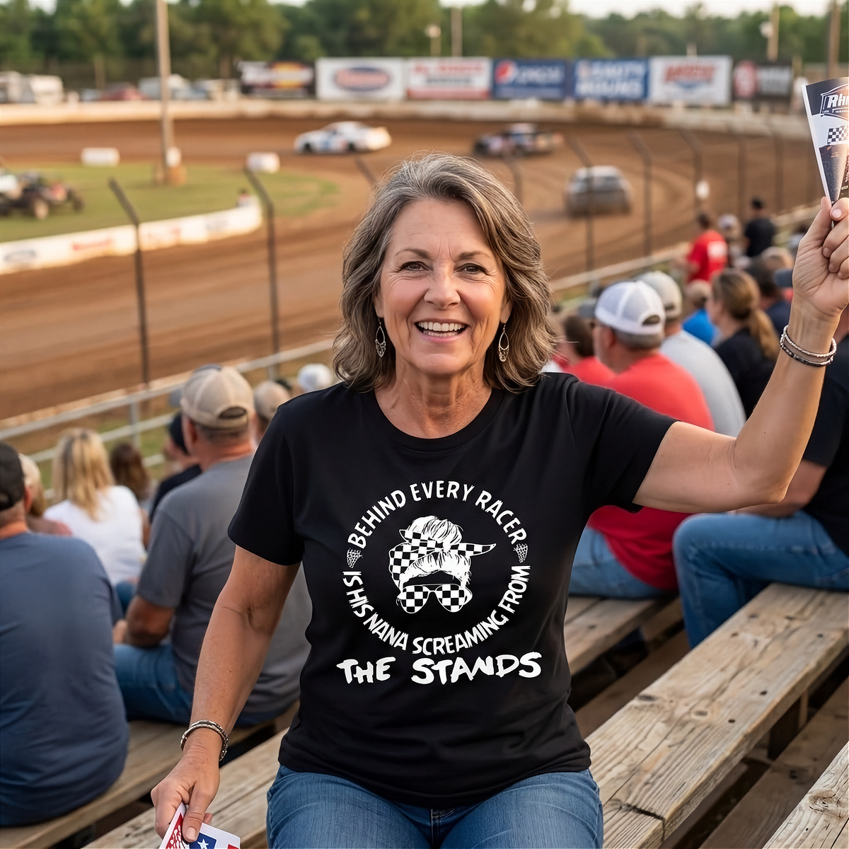 A woman stands on a racetrack bleachers wearing a black shirt with a white graphic print that reads 'BEHIND EVERY RACER IS HIS NANA SCREAMING FROM THE STANDS' with a checkerboard flag design. Race cars are visible in the background