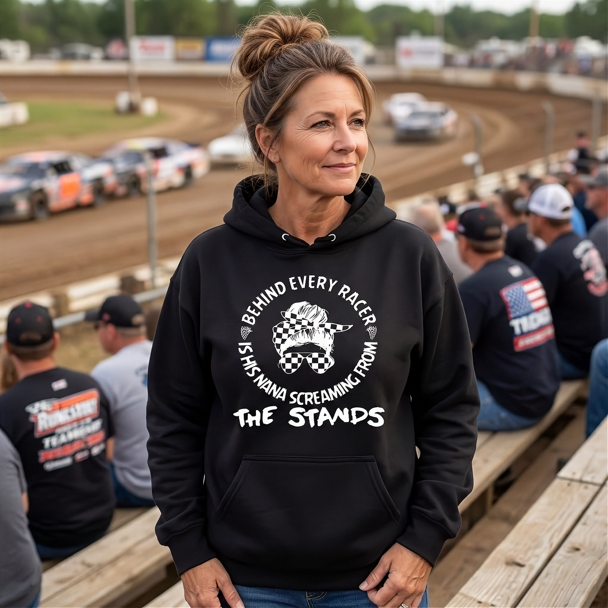 A woman stands on a racetrack bleachers wearing a black hoodie with a white graphic print that reads 'BEHIND EVERY RACER IS HIS NANA SCREAMING FROM THE STANDS' with a checkerboard flag design. Race cars are visible in the background