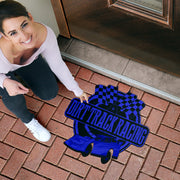 A woman kneels on a brick driveway next to a door, pointing to a decorative doormat featuring a blue and black dirt track racing design with a race car and checkered flag.