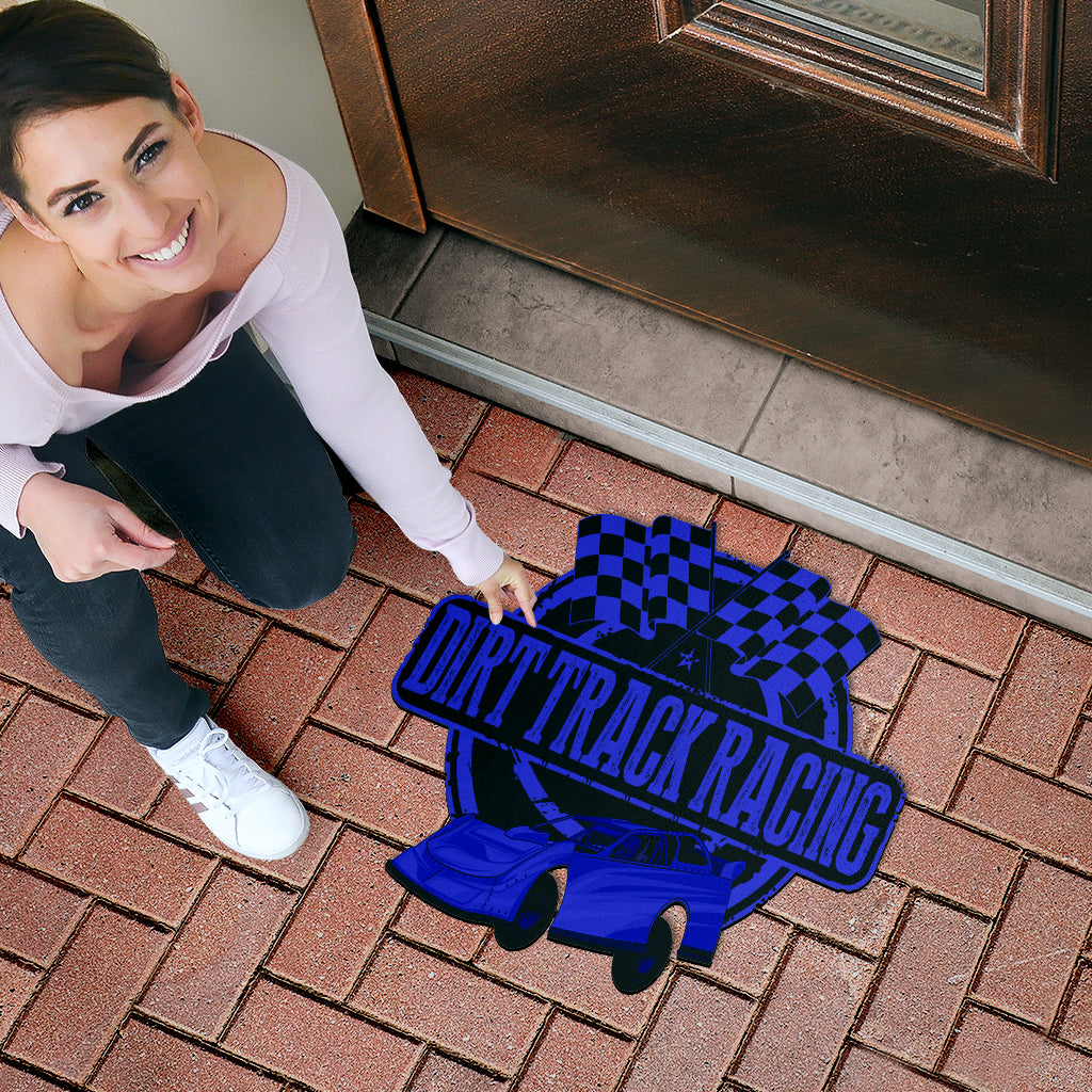 A woman kneels on a brick driveway next to a door, pointing to a decorative doormat featuring a blue and black dirt track racing design with a race car and checkered flag.