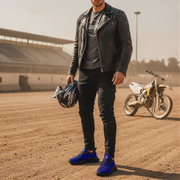 Man in leather jacket holding a helmet and blue checkered sneakers on a dirt track with a motorcycle in the background