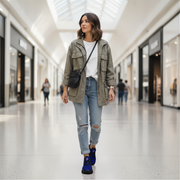 Woman walking in a modern mall wearing an olive green jacket, white shirt, ripped jeans, and blue checkered shoes.