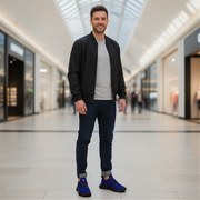 Man standing in a modern mall wearing a black jacket, gray shirt, blue jeans, and blue sneakers.
