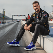 Man sitting on a race track wearing a black racing suit and blue checkered sneakers.