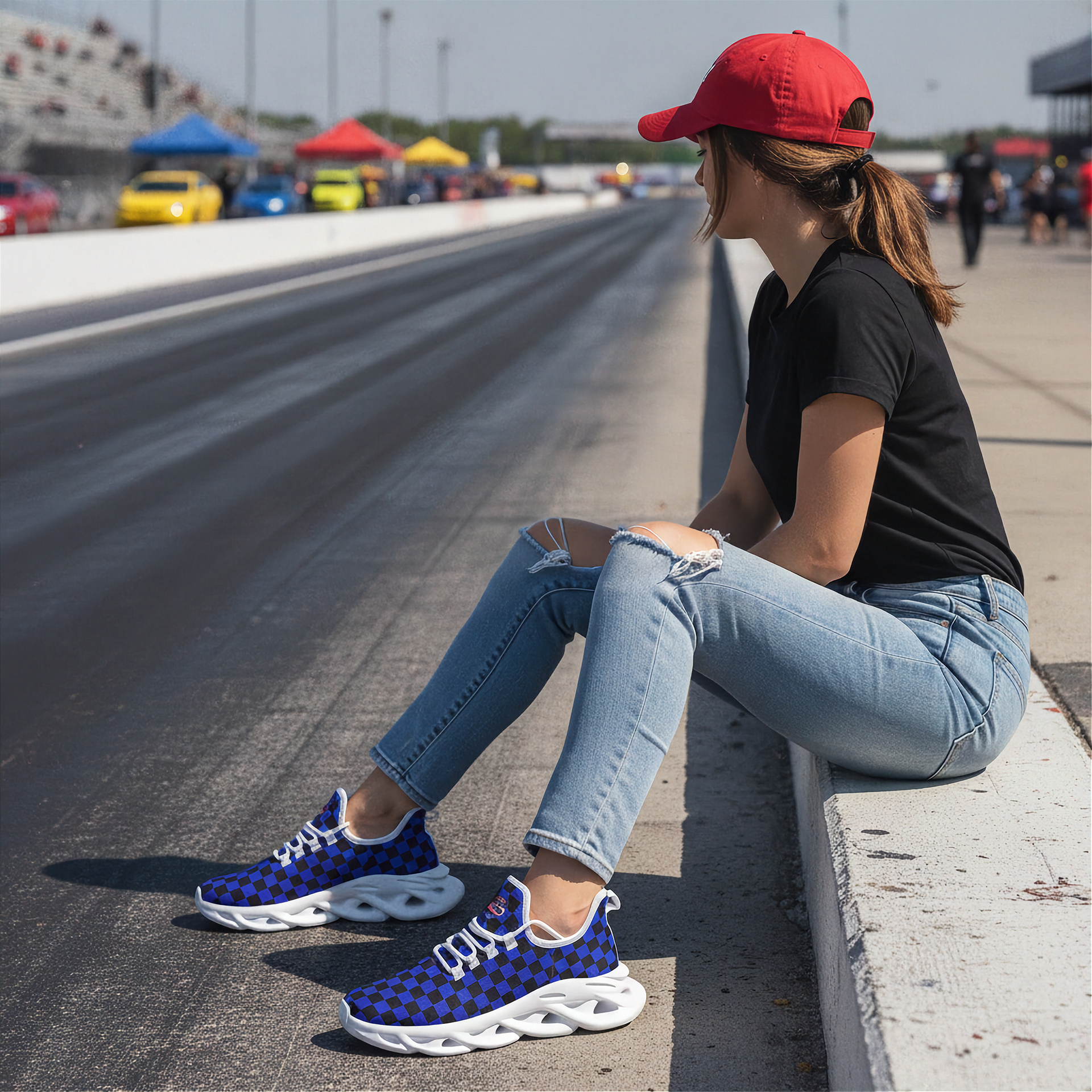 Woman sitting on a race track wearing blue sneakers with white soles, black shirt, and red cap.