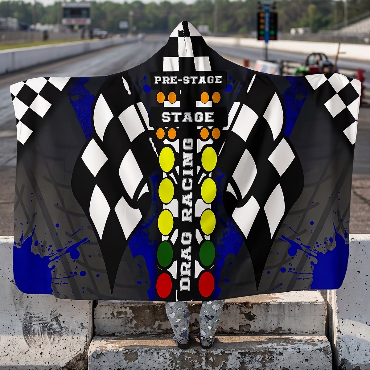 A woman standing at a drag race track wearing a hooded blanket with a big drag racing tree logo.
