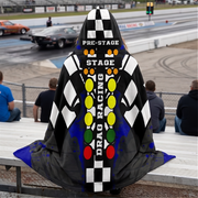 A woman sitting at the stands at a drag race track wearing a hooded blanket with a drag racing theme. The garment features a black-and-white checkered pattern, blue splatter accents, and a central vertical graphic with the words 'PRE-STAGE', 'STAGE', and 'DRAG RACING' over a red, yellow, green, and orange light-tower motif.
