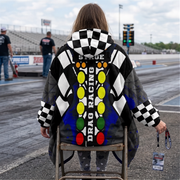 A woman sitting on a chair at a drag race track wearing a hooded blanket with a drag racing theme. The garment features a black-and-white checkered pattern, blue splatter accents, and a central vertical graphic with the words 'PRE-STAGE', 'STAGE', and 'DRAG RACING' over a red, yellow, green, and orange light-tower motif.