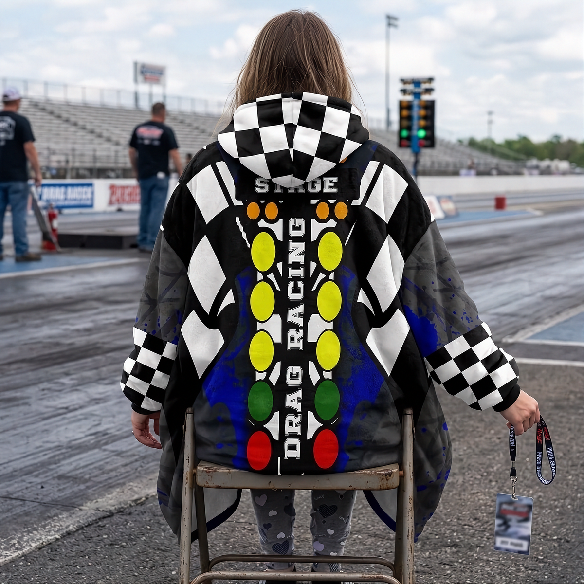 A woman sitting on a chair at a drag race track wearing a hooded blanket with a drag racing theme. The garment features a black-and-white checkered pattern, blue splatter accents, and a central vertical graphic with the words 'PRE-STAGE', 'STAGE', and 'DRAG RACING' over a red, yellow, green, and orange light-tower motif.