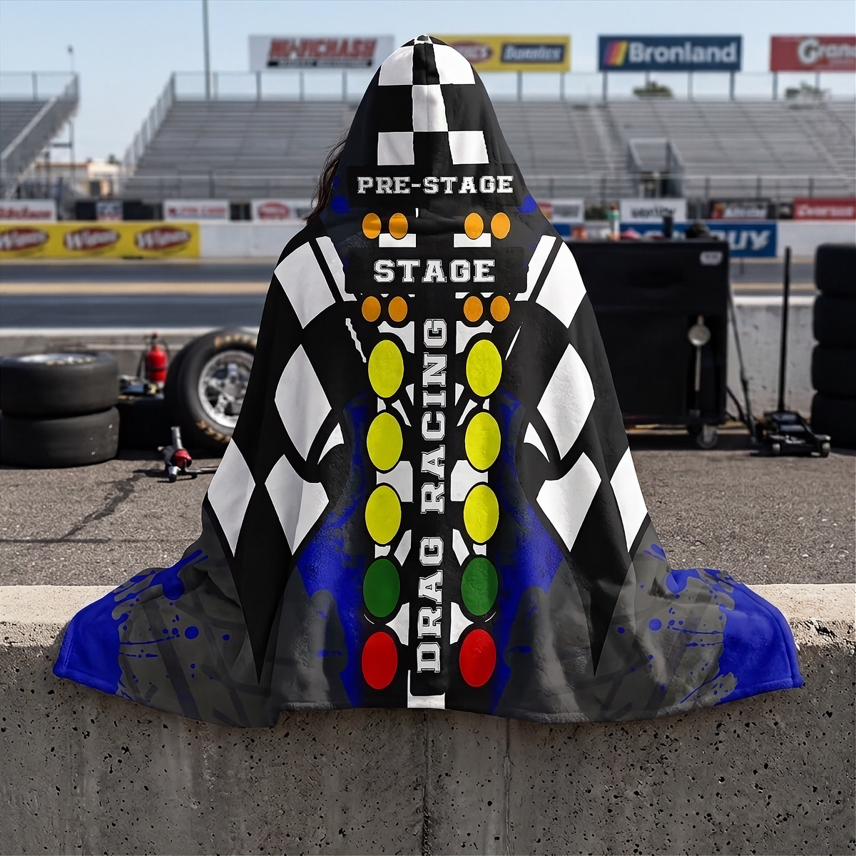A woman sitting on a concreate wall at a drag race track wearing a hooded blanket with a drag racing theme. The garment features a black-and-white checkered pattern, blue splatter accents, and a central vertical graphic with the words 'PRE-STAGE', 'STAGE', and 'DRAG RACING' over a red, yellow, green, and orange light-tower motif.