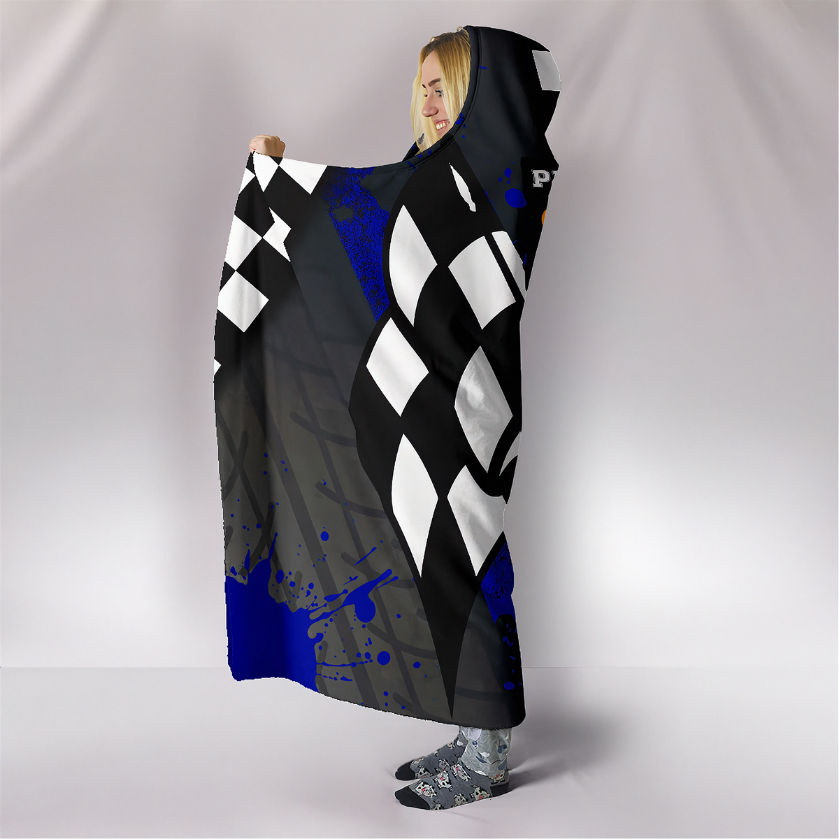 A person wearing a hooded blanket or throw with a black, white, and gray harlequin diamond pattern and blue paint splatter accents, standing against a light gray studio backdrop.