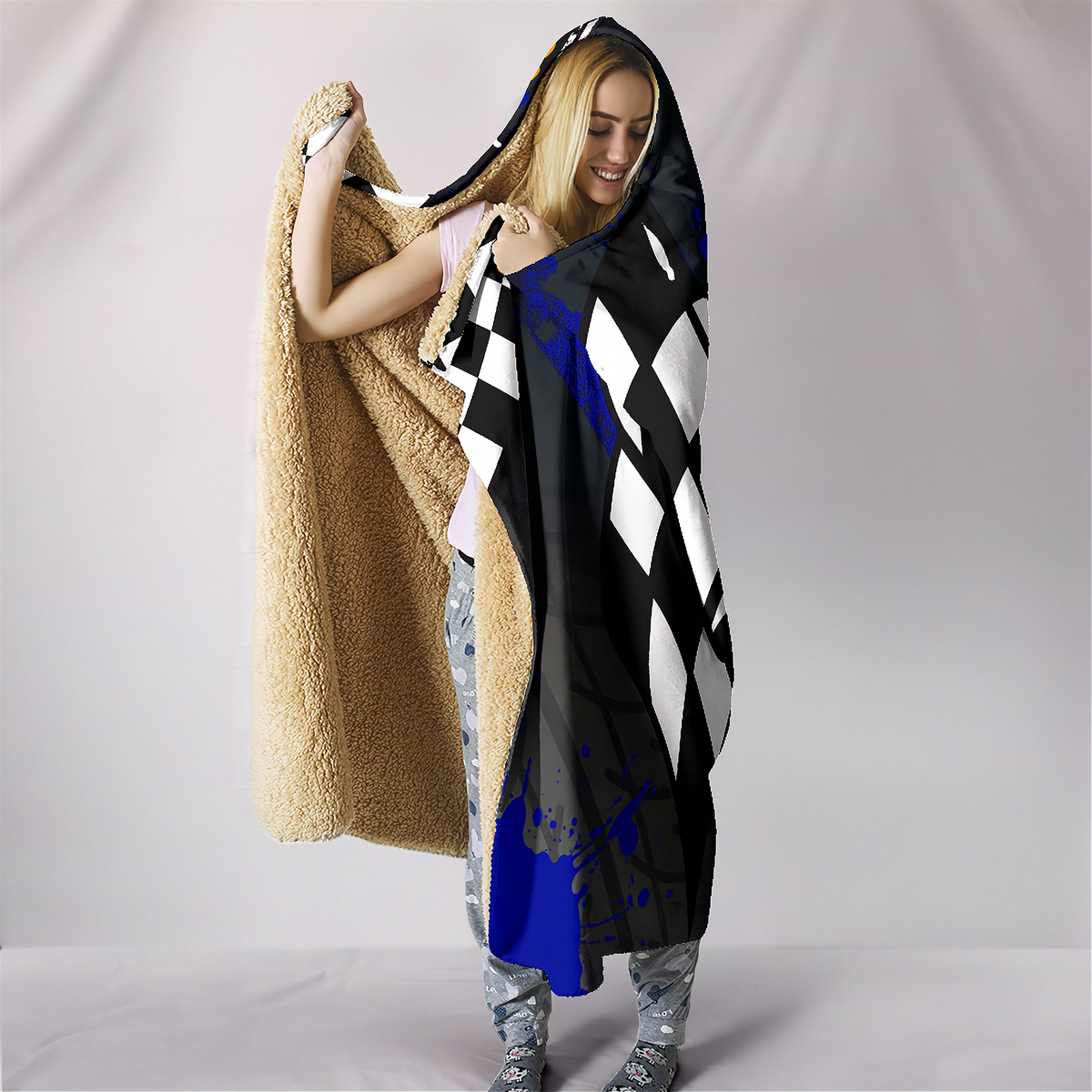 A person wrapped in a hooded fleece blanket with a black-and-white harlequin pattern, blue paint splatter accents, and a tan sherpa lining, standing against a light gray studio backdrop.