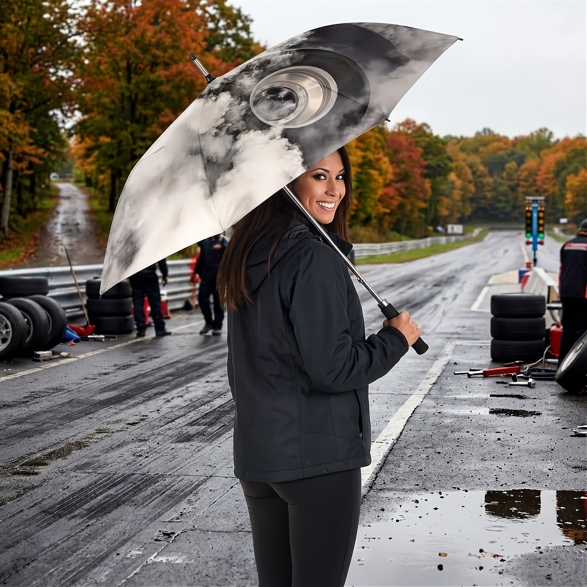 A woman on a wet racetrack holds a large open umbrella with a black-and-white cloud graphic. She wears a dark rain jacket and pants, with trees in autumn colors and race track equipment visible in the background.