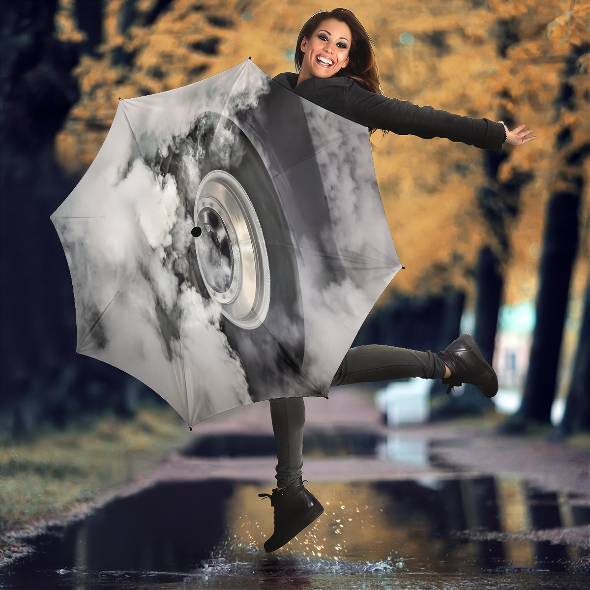 A woman jumps in a puddle outdoors while holding a large open umbrella printed with a black-and-white tire and cloud design. Trees with yellow autumn foliage are blurred in the background.