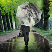A person walking in the rain along a tree-lined path, holding a large umbrella printed with a dramatic black-and-white tire and cloud design.