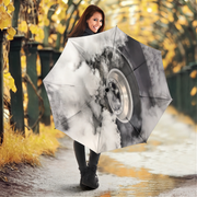 A woman holding a large open umbrella in an outdoor autumn setting. The umbrella features a printed design of a silver car wheel surrounded by white and gray clouds.