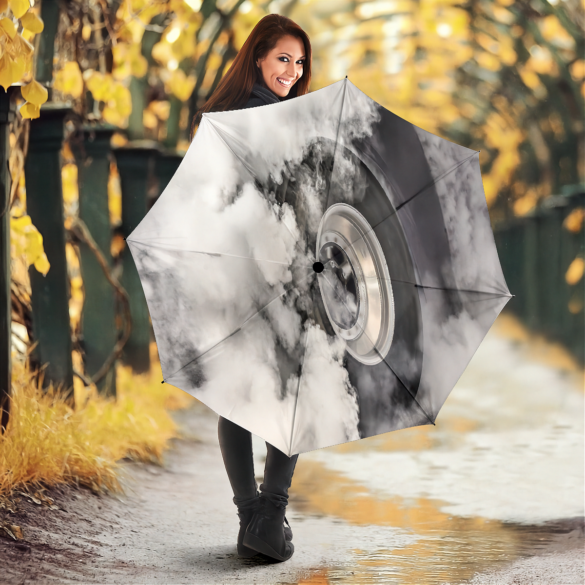 A woman holding a large open umbrella in an outdoor autumn setting. The umbrella features a printed design of a silver car wheel surrounded by white and gray clouds.