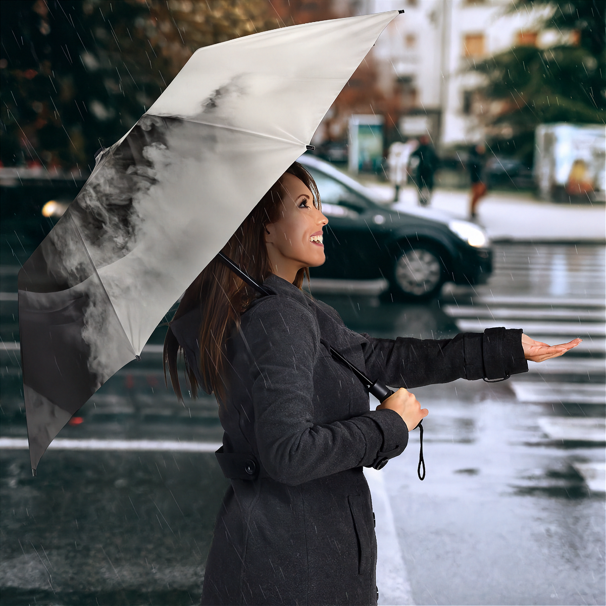A woman in a dark gray coat stands in the rain holding a large open umbrella with a black and white smoky graphic print. A wet city street, blurred car, and pedestrians are visible in the background.