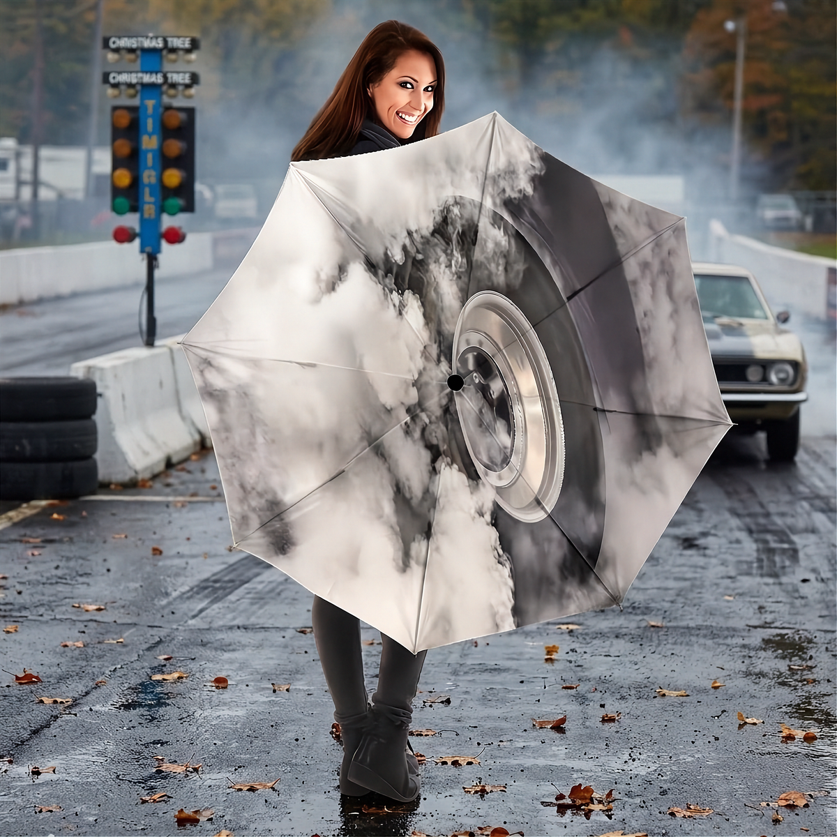 A woman holding a large open umbrella in an outdoor street setting. The umbrella features a dramatic printed design of a car wheel partially covered by swirling white and gray smoke.