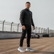 Man in black jacket and pants and checkered shoes standing on a race track with a car in the background