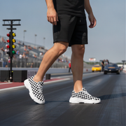 Man wearing black checkered sneakers on a racetrack with cars and traffic lights in the background.