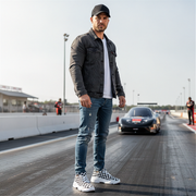 Man standing on a race track wearing a black jacket and jeans and checkered shoes with a car in the background.