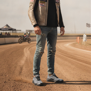 Person standing on a dirt track wearing a jacket with patches, jeans, and checkered shoes.