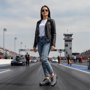 Woman in leather jacket and jeans  and checkered shoes standing on a race track with cars and a control tower in the background.