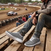 Man wearing black-and-white checkered sneakers with white soles while seated in dirt track grandstands.