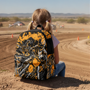 A child wearing a black and orange graphic backpack sits on a dirt track in a desert landscape, viewed from behind.