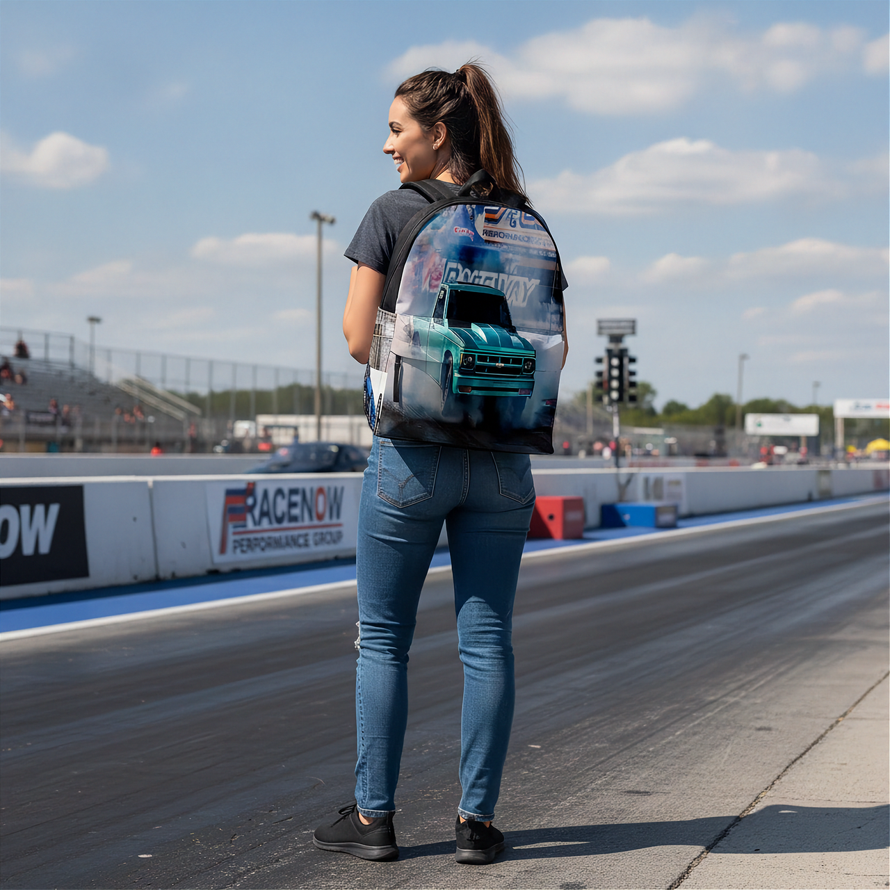 A woman stands on a drag racing track wearing a custom printed backpack with a 3D-effect graphic of a teal race car. The background shows the racetrack, bleachers, and blue sky.