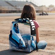 A child sits on a racetrack wearing a backpack printed with a blue and white dragster and the word 'DRAG RACING' on the side, with the racetrack and grandstands blurred in the background.
