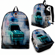A backpack shown from multiple angles on a plain white background, featuring a wraparound drag racing truck graphic with smoke, track signage, and a turquoise pickup truck. No person, animal, or real environment is present, so this is Product/Packaging.