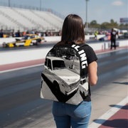 A woman standing on a racetrack wearing a black backpack with a large black-and-white vintage car graphic and bold white vertical lettering on the side, paired with a black t-shirt and blue jeans.