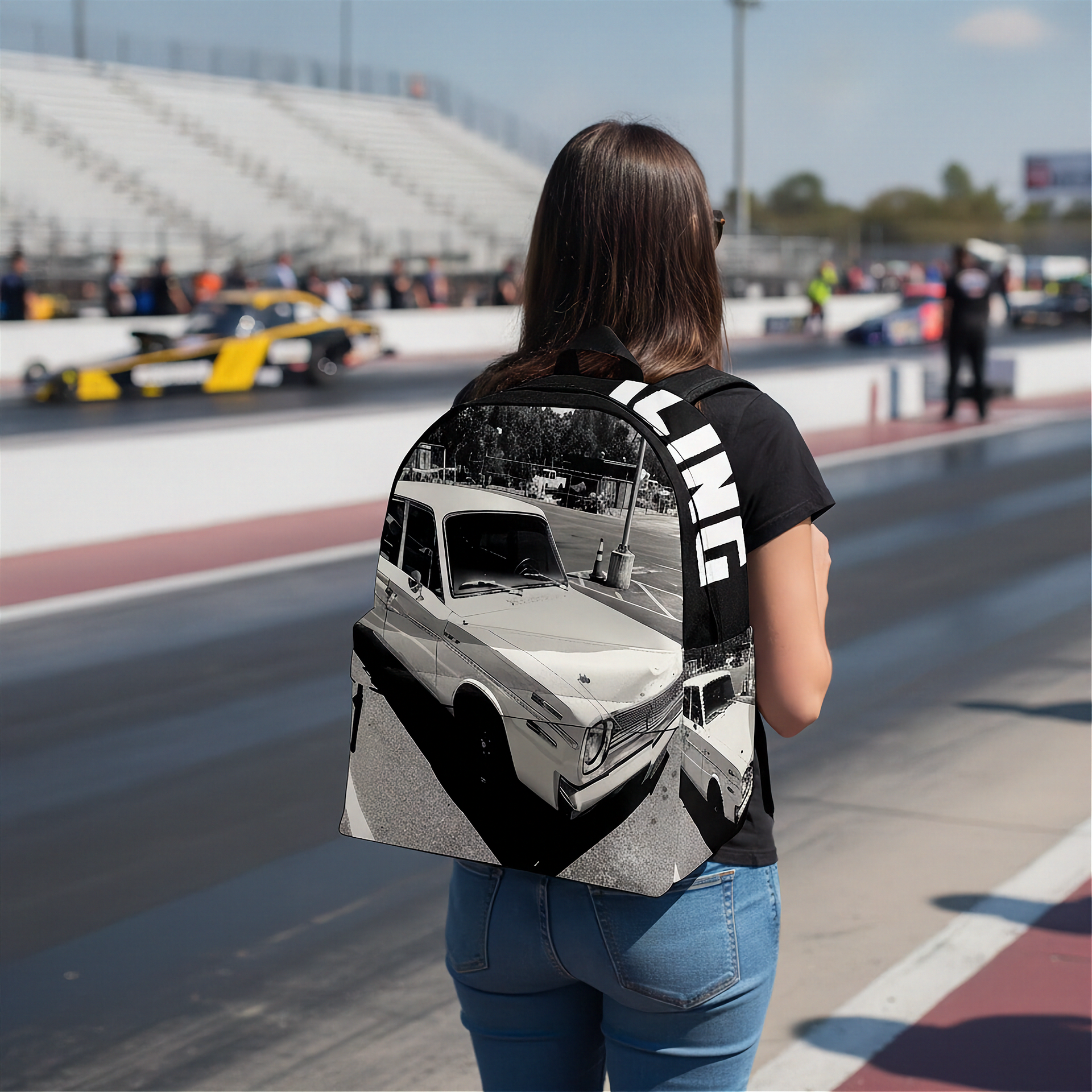 A woman standing on a racetrack wearing a black backpack with a large black-and-white vintage car graphic and bold white vertical lettering on the side, paired with a black t-shirt and blue jeans.