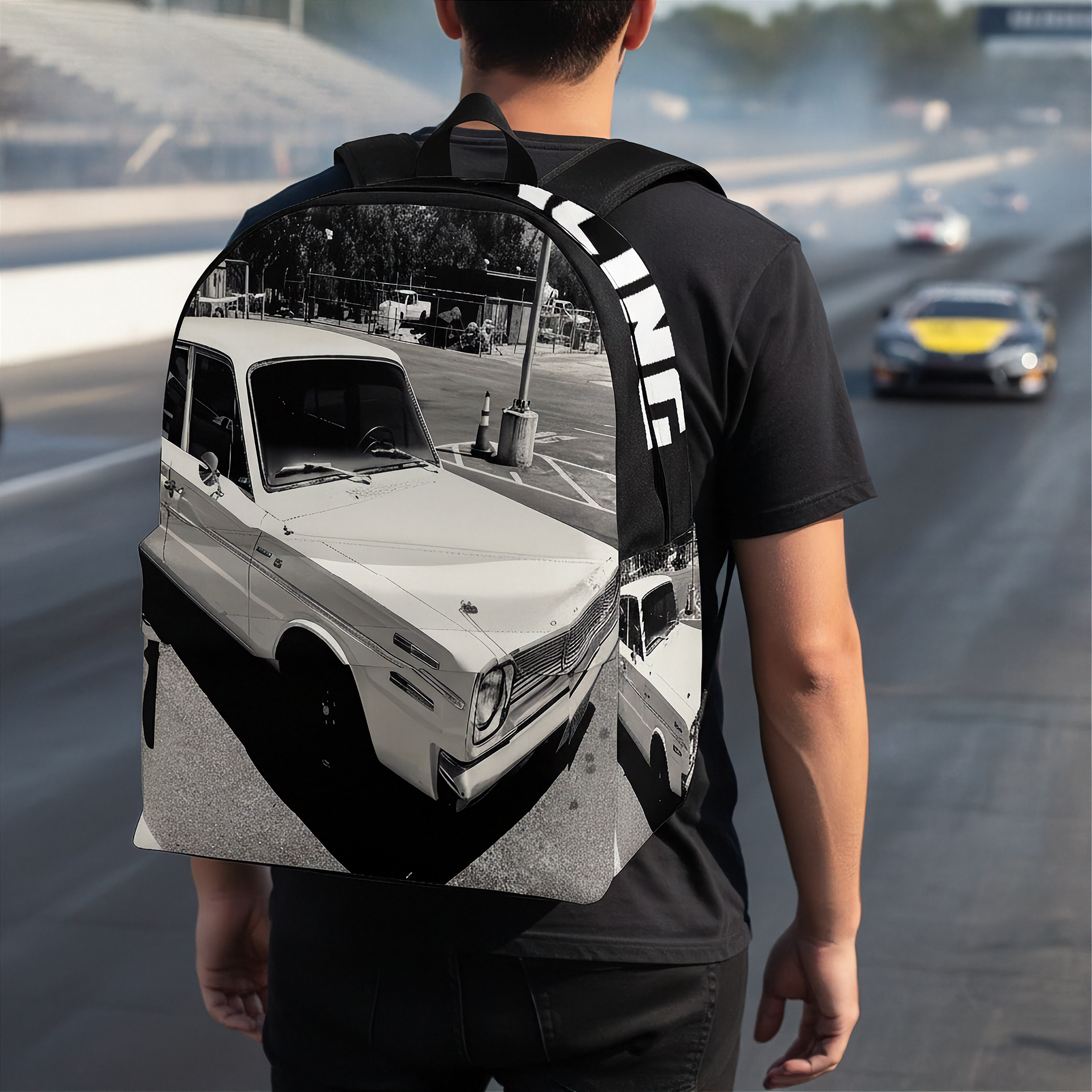 A man wearing a black t-shirt and black pants, viewed from behind, carrying a backpack on their shoulders. The backpack features a large graphic print of a vintage white car in a black-and-white photo style, with the word 'FINITE' printed vertically in white on the black shoulder strap.