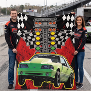 A man and a woman standing on a racetrack holding up a large printed blanket featuring a green drag racing car, checkered flags, traffic lights, and the words 'PRE STAGE STAGE DRAG RACING'.