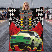 A blond woman stands on a racetrack holding up a large printed blanket featuring a green drag racing car, checkered flags, traffic lights, and the words 'PRE STAGE STAGE DRAG RACING'.