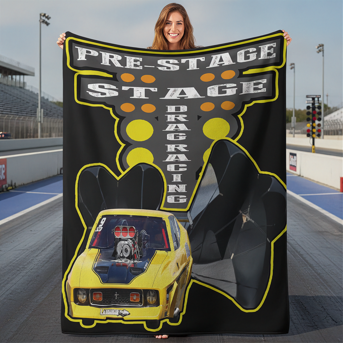 A woman stands on a drag racing track holding up a large black fleece blanket or throw. The blanket features a yellow and black graphic design with the text 'PRE-STAGE STAGE DRAGRACING' and an illustration of a yellow race car.