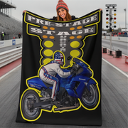 A woman wearing a red knit beanie holds up a black fleece blanket featuring a graphic of a rider on a blue Kawasaki motorcycle with 'PRE-STAGE STAGE' text, set against a racetrack background.