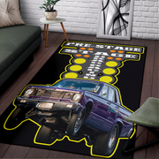 A rectangular area rug featuring a purple classic muscle car and the words 'PRE-STAGE STAGE FIGHT' in bold yellow, black, and white lettering, shown on a dark wood floor in a living room setting.