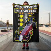 Person holding a 'Pre-Stage Stage Parking' blanket with a race car design on a race track.